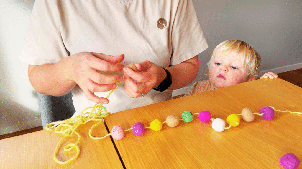 Toddler looking on at colorful felted balls are strung up into a garland