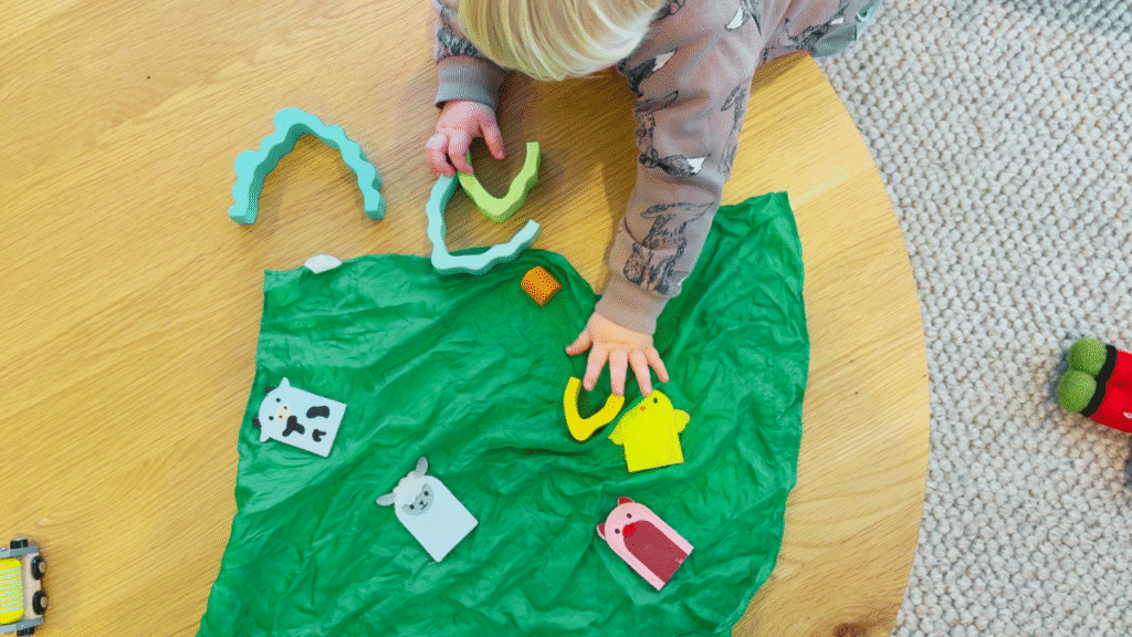 Toddler engaging in independent play with Waldorf-inspired toys, including felt farm animal finger puppets, rainbow arches, and a green silk play mat on a wooden table.