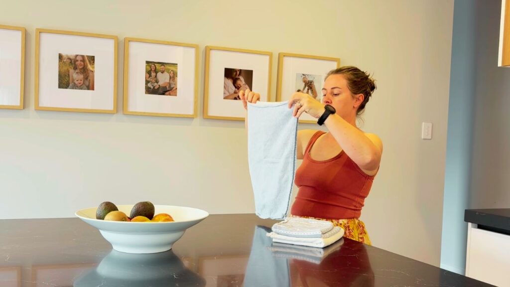 Woman folding kitchen towels on a dark countertop with family photos on the wall — calm daily rhythm as part of a kitchen refresh for a new year of flow.