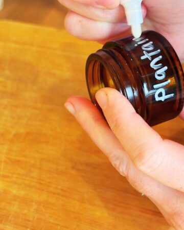Handwriting a label that says “Plantain Salve” on a small amber glass jar for natural first-aid balm.