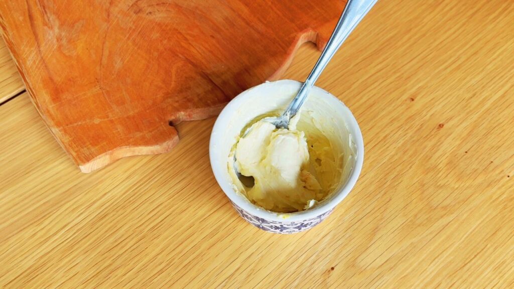 Small ceramic bowl filled with leftover cream cheese frosting beside a wooden chopping board on a kitchen table.
