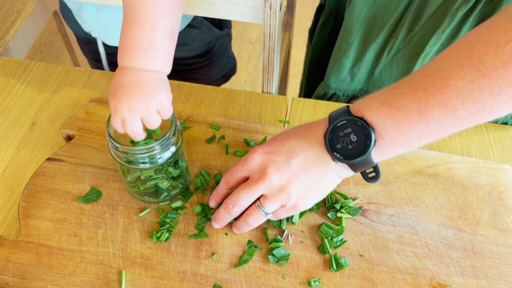 Mother and toddler chopping plantain leaves and filling a small jar together — making a natural herbal balm.