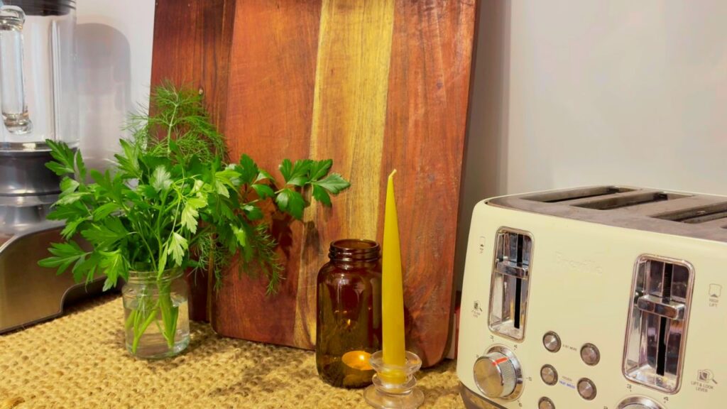 A calm kitchen counter with fresh herbs in a jar, beeswax candle, amber glass bottle, wooden chopping board, and cream toaster — natural tones reflecting a kitchen refresh for a new year of flow.