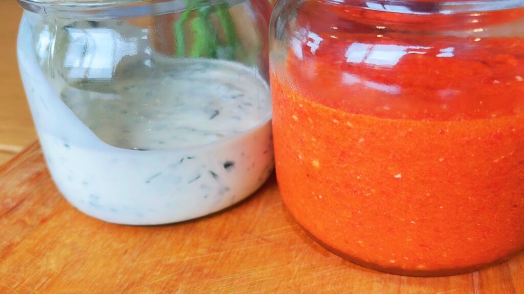 A glass jar of creamy homemade ranch dressing beside a jar of bright red fermented sriracha on a wooden board — both made from real, seed-oil-free ingredients that support a healthy gut microbiome.