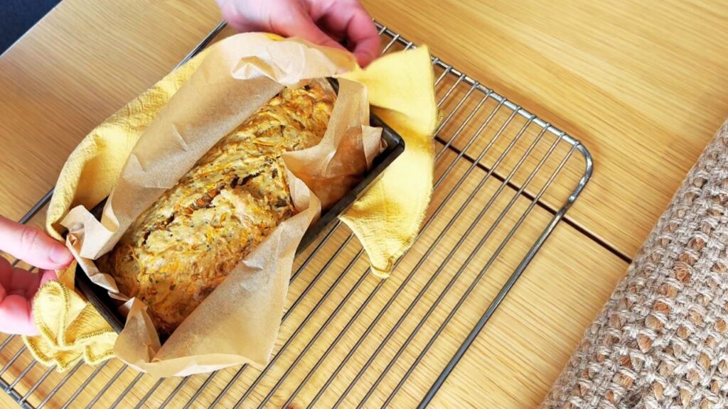 Freshly baked gut friendly carrot cake in a loaf tin lined with parchment paper, cooling on a wire rack with a yellow tea towel.