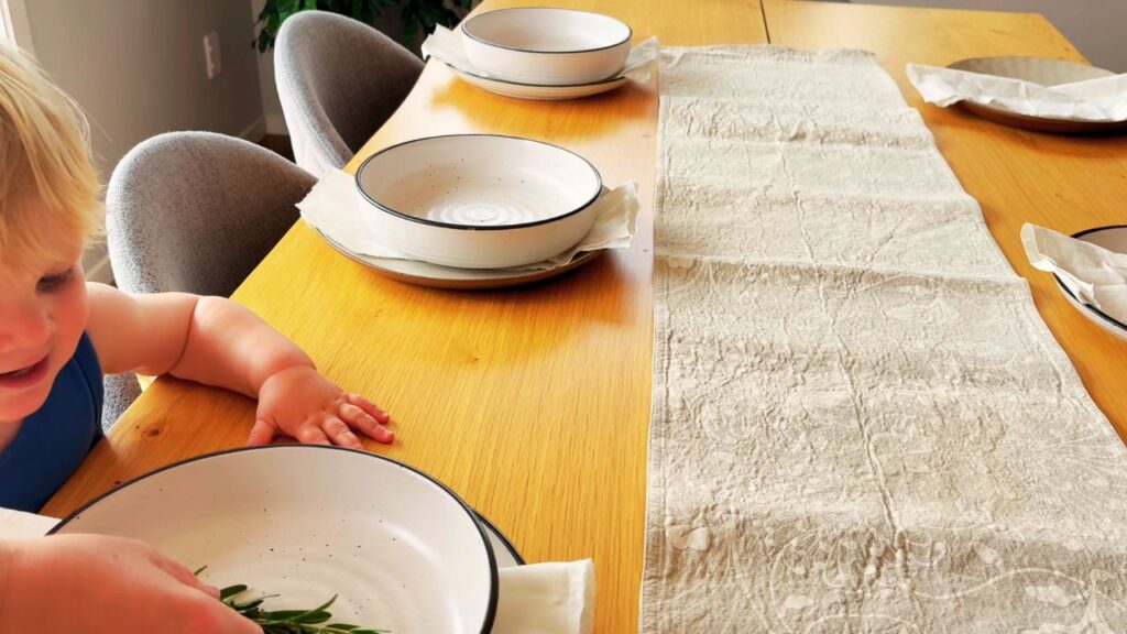 A toddler reaches toward a bowl with sprigs of rosemary on a wooden table, showing the warmth and togetherness of calm family dinners.