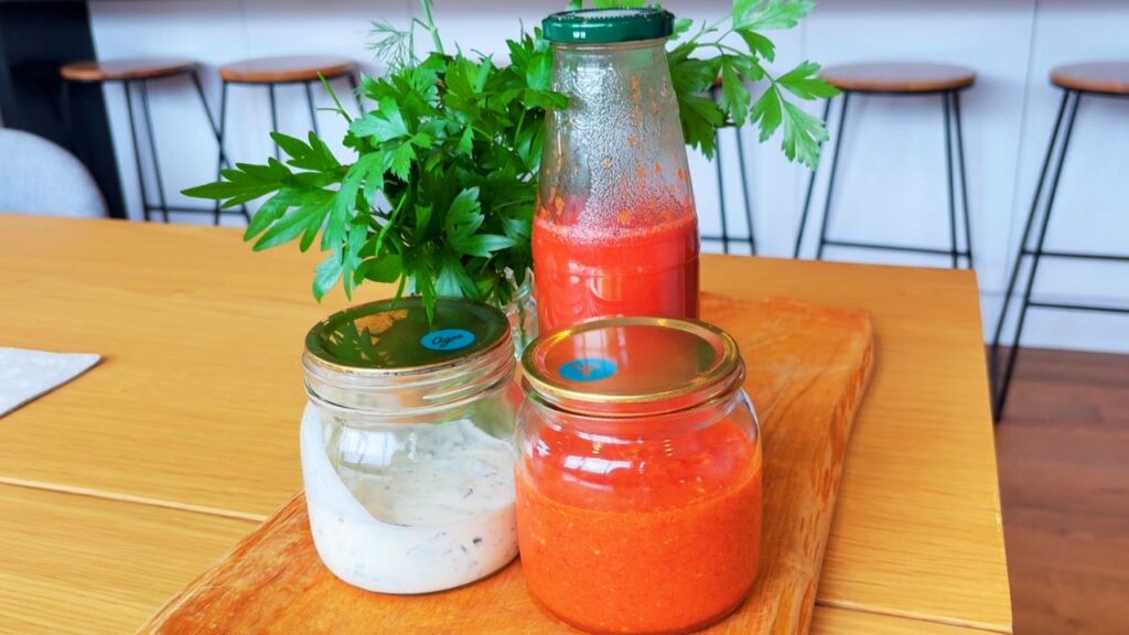 Three jars of homemade seed-oil-free condiments — ketchup, ranch, and sriracha — displayed on a wooden board with fresh parsley in the background, capturing a wholesome, gut-healthy kitchen scene.