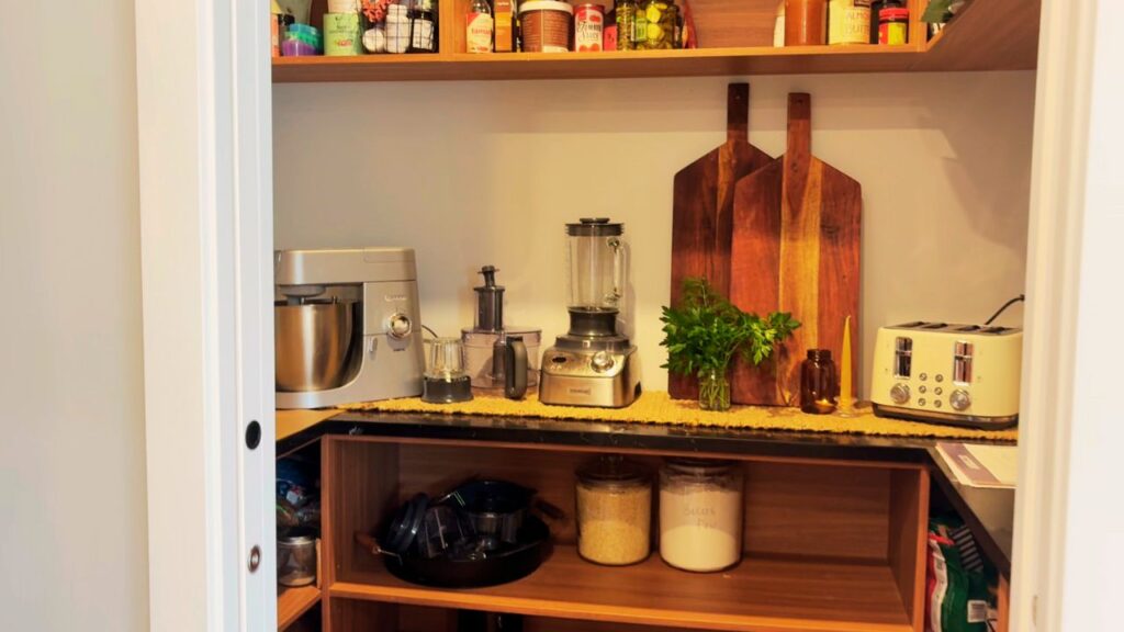 Wide view of a minimalist pantry with wooden shelves, chopping boards, appliances, and neatly arranged jars — a family kitchen refreshed for more calm and flow