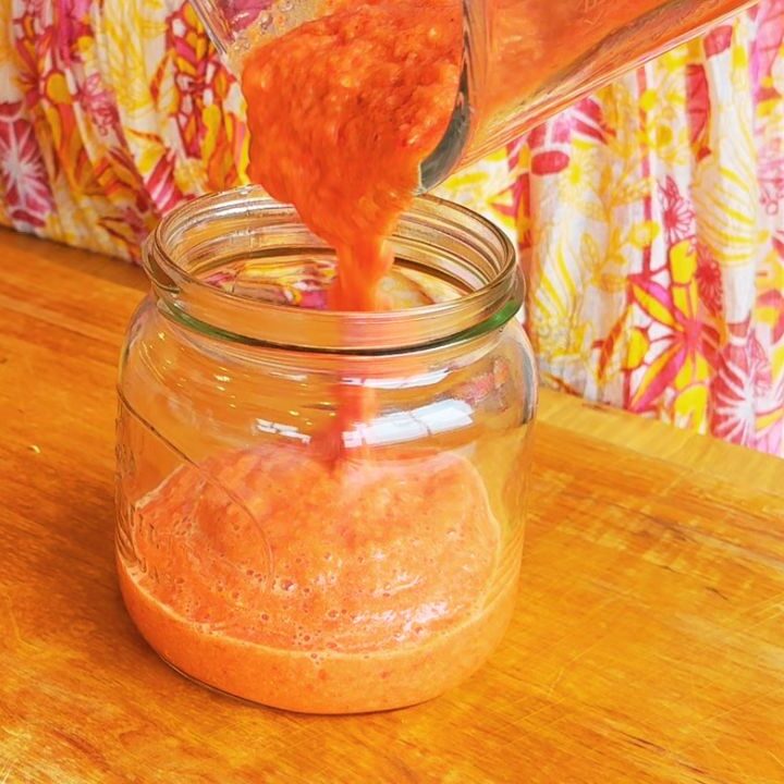 Pouring freshly blended red sriracha into a glass jar on a wooden surface, showing the thick, vibrant texture of this fermented, gut-microbiome-supporting condiment.