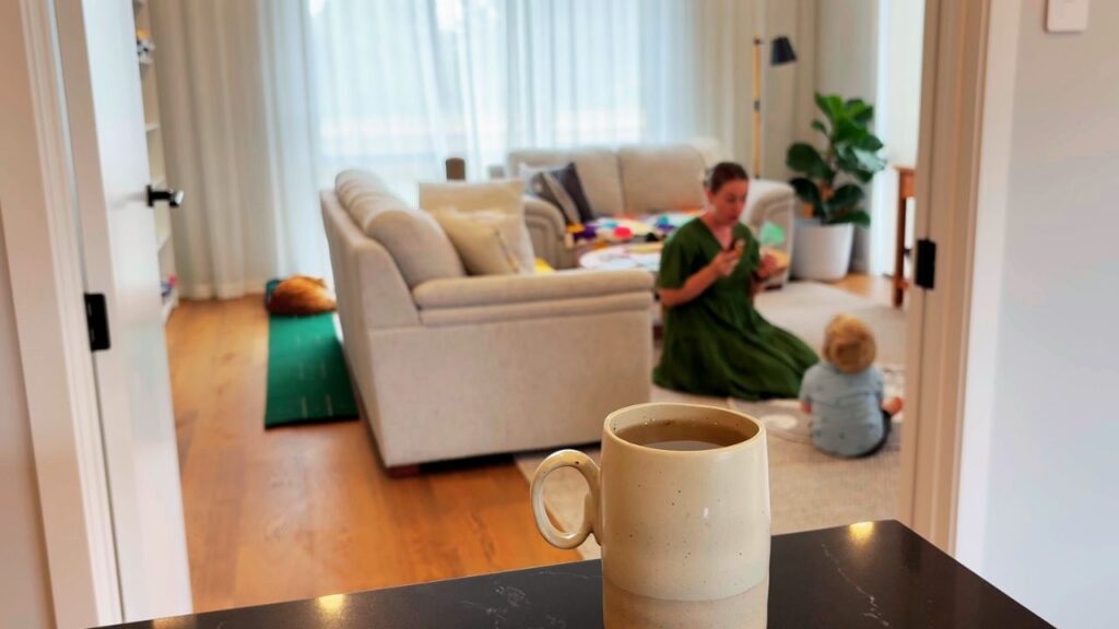 A warm mug of coffee on the counter with family storytelling and play unfolding in the sun-filled living room behind.