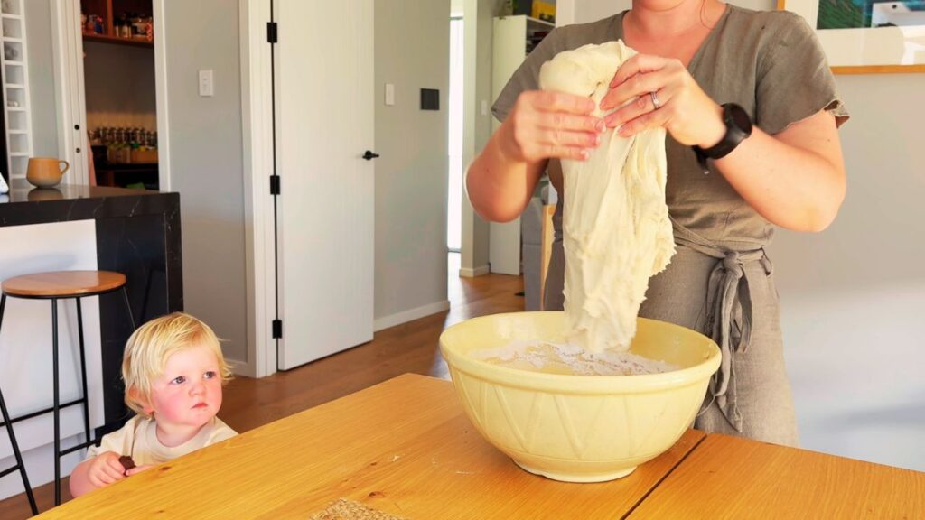 Parent stretching sourdough dough at the kitchen table with toddler watching—making real-food bread without vegetable oil.