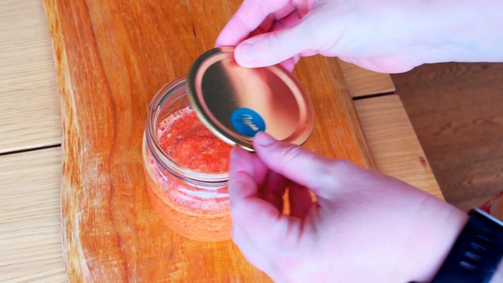 Hands sealing a jar of freshly made fermented sriracha with a golden lid, representing real food condiments made without seed oils or refined sugar.