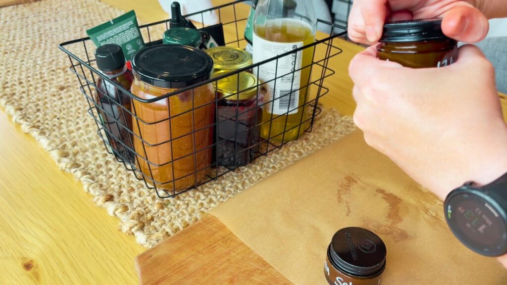 Hands opening a small brown jar of homemade plantain salve beside a basket of natural oils and herbal ingredients.