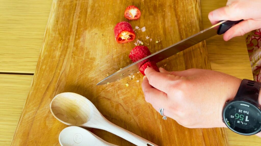 Slicing red chili peppers on a wooden board with coarse salt and wooden spoons nearby — the simple start of a fermented, seed-oil-free sriracha.