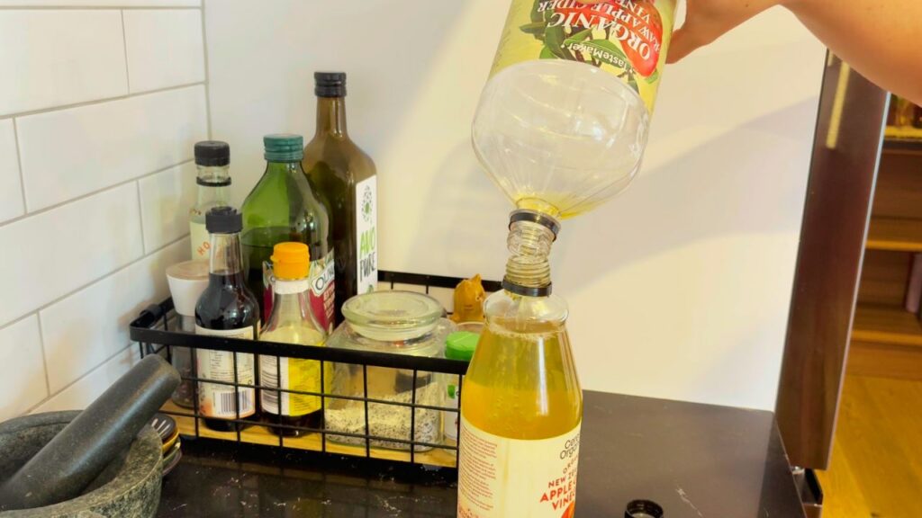 Refilling a bottle of organic apple cider vinegar beside olive oil and avocado oil — a natural kitchen refresh moment focused on real-food simplicity