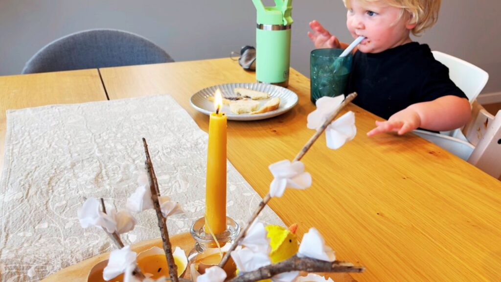Toddler drinking from a cup at the family table with a lit beeswax candle and simple seasonal centerpiece in the foreground.