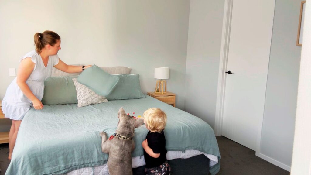 Mother making the bed while her toddler and family dog stand nearby wanting to help as part of a calm home rhythm.
