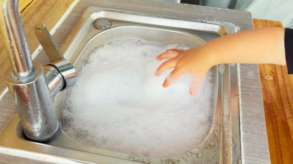 Child’s hand playing with soap bubbles in the sink of an outdoor mud kitchen.