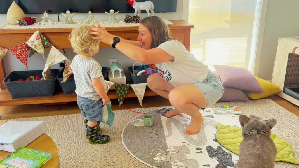 A playful moment in the living room as a mother crouches to engage with her toddler, surrounded by Christmas bunting and cozy toys.