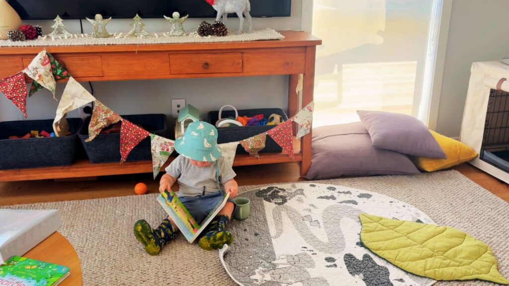 A toddler sitting on the living room floor reading a book independently, with Christmas bunting and seasonal decorations on the cabinet behind.