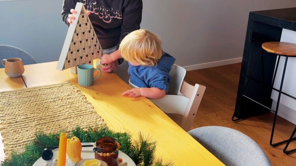 A parent and toddler opening the wooden Advent calendar together at the dining table, with warm morning light and a simple Christmas centerpiece.