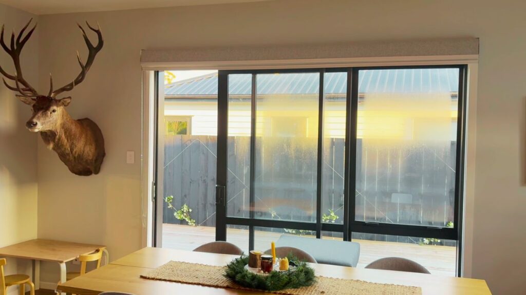 A dining room filled with golden sunset light, a Christmas wreath centerpiece on the table, and a mounted deer head on the wall, with large glass doors opening onto the deck outside.