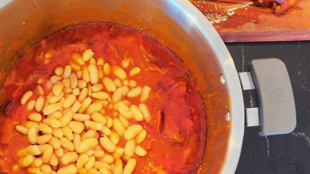 Adding cannellini beans to slow-cooked ham hock baked beans during a real-food breakfast prep for the week.