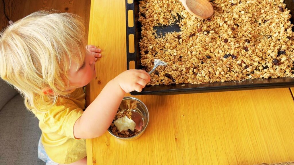 Toddler serving himself homemade granola with yoghurt at the table, part of our simple gut-friendly breakfast rhythm.