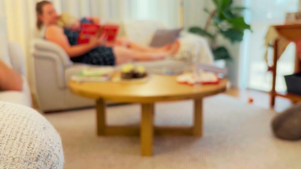 A cozy living room scene with a mother and toddler reading together on the couch, soft summer light coming through the curtains and a relaxed, slow-living atmosphere.
