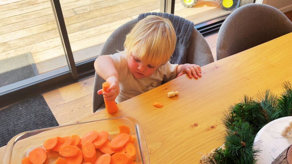 Toddler exploring carrot slices at the table while helping prepare simple holiday side dishes.