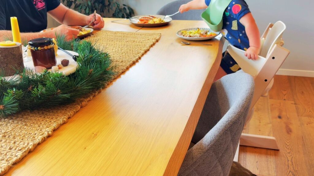 A family sitting at the dining table for a simple meal, with a toddler in a high chair and a handmade Christmas wreath centerpiece adding a festive touch.