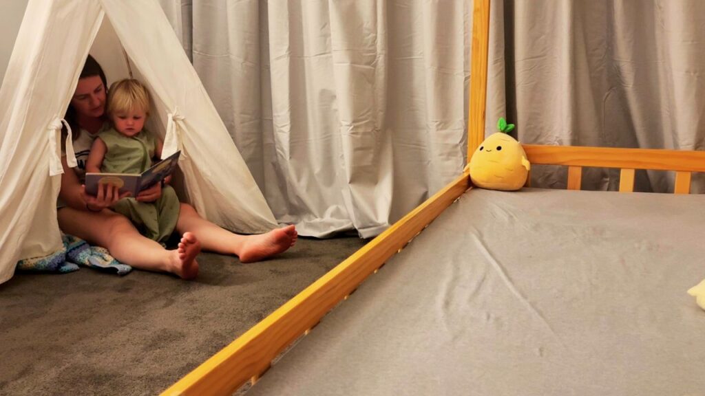 A toddler lying on the bedroom floor getting ready for bed as a parent helps with pajamas, with a storybook and cozy play tent nearby.